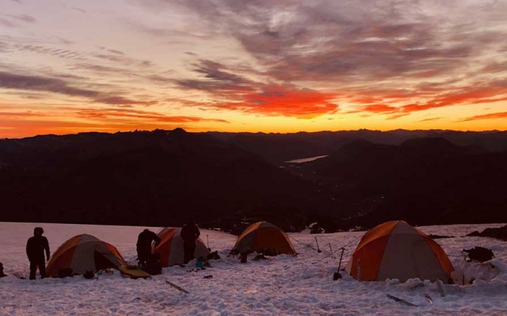 Four tents rest in the snow of a campsite. In the background, the sky appears in vibrant yellow and orange, as the sun rises or sets.